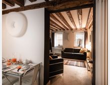 Interno di un appartamento con soggiorno e zona pranzo, caratterizzato da soffitto con travi in legno a vista e pareti bianche. Nel soggiorno ci sono divani scuri, tappeto colorato e due finestre con inferriate e tende, mentre in primo piano si vede un tavolo apparecchiato in vetro.