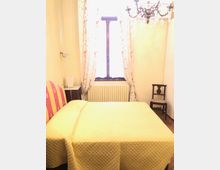 Camera da letto semplice con letto matrimoniale e copriletto chiaro, una finestra centrale con tende leggere e un radiatore sotto. Sulla destra c’è una sedia in legno e in alto un lampadario, con pavimento in legno e pareti chiare.