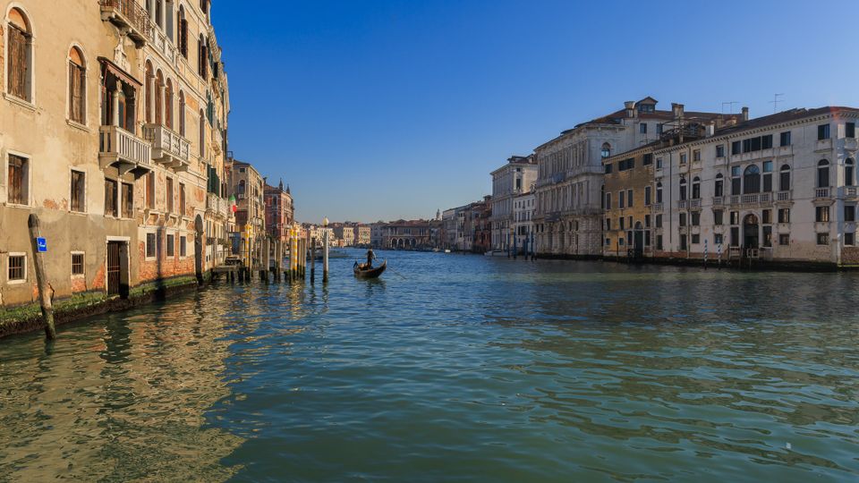 Ampio canale cittadino fiancheggiato da palazzi storici con facciate chiare, finestre ad arco e balconi, con pali d’ormeggio lungo le rive. Acqua calma al centro del canale sotto cielo sereno, con una gondola in lontananza.