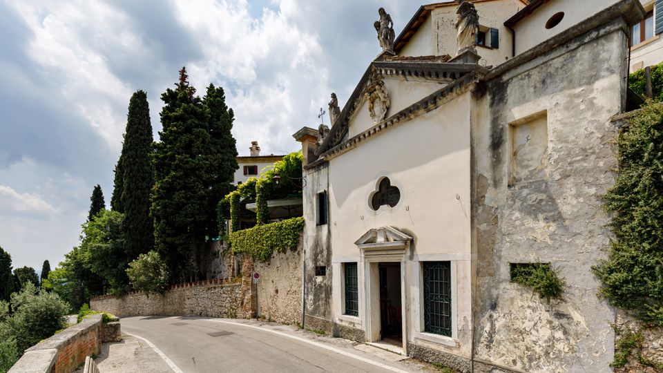 Piccola chiesa storica intonacata bianca lungo una strada curva, con facciata a timpano decorata da statue e una finestra trilobata sopra l’ingresso. Accanto ci sono muri in pietra coperti di vegetazione e alti cipressi, con cielo parzialmente nuvoloso sullo sfondo.