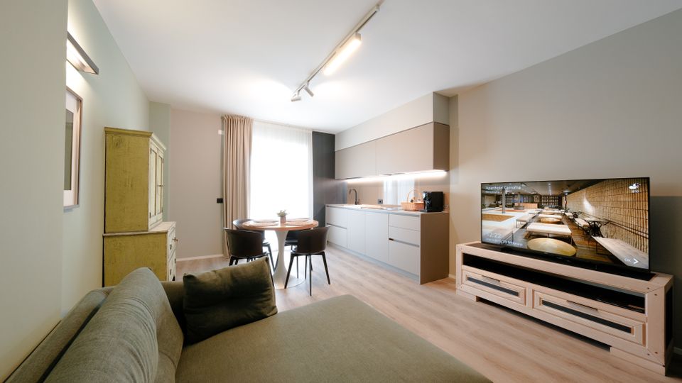 Soggiorno open space moderno con cucina lineare a parete, tavolo rotondo con sedie vicino a una grande finestra con tende e divano in primo piano. A destra ci sono un mobile TV basso con televisore, mentre a sinistra un armadio in stile rustico; pavimento in legno chiaro e illuminazione a binario sul soffitto.