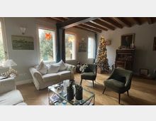 Soggiorno rustico con travi in legno a vista, divano chiaro e due poltrone scure attorno a un tavolino in vetro. Sullo sfondo si vedono una finestra con luce naturale, un camino e un albero di Natale decorato vicino alla parete.