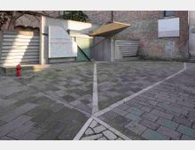 Piccolo cortile pavimentato in pietra con linee bianche geometriche, affiancato da un edificio in mattoni e da pareti in cemento con pannelli e portoni a lamelle. Sono presenti una pensilina sporgente e un idrante rosso vicino al muro.