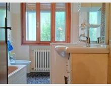 Bagno domestico luminoso con grande finestra a tre ante con telaio in legno e radiatore sotto. In primo piano si vedono il lavabo con mobile e specchio, mentre a sinistra è presente una vasca e il pavimento è rivestito con piastrelle scure.