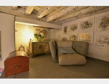 Piccolo salotto mansardato con soffitto a travi di legno a vista e pareti chiare. Nell’angolo ci sono una poltrona imbottita, una cassapanca antica e un mobile decorato, con parete impreziosita da quadri floreali e illuminazione soffusa.