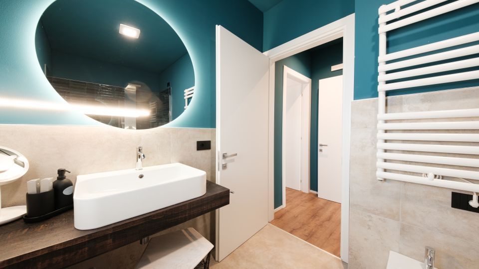 Bagno moderno con pareti blu petrolio e rivestimento in piastrelle grigie, lavabo rettangolare su mensola in legno scuro e grande specchio rotondo retroilluminato. A destra si vede un termoarredo bianco e, sul fondo, la porta aperta verso un corridoio con pavimento in legno.