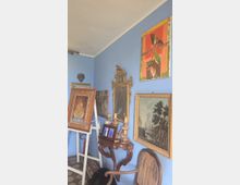 Angolo di una stanza con pareti azzurre, arredato con un cavalletto che regge un quadro, un tavolino intagliato con candelieri e piccoli oggetti, e uno specchio antico decorato. Alle pareti sono appesi dipinti incorniciati, una maschera e un quadro moderno, con pavimento a piastrelle geometriche e una poltrona in primo piano.