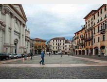 Ampia piazza lastricata in un centro storico, con una grande chiesa in stile classico sul lato sinistro e una fila di palazzi colorati con portici e balconi sul lato destro. Sullo sfondo si vedono altri edifici e un cielo nuvoloso, con auto parcheggiate lungo il margine della piazza.