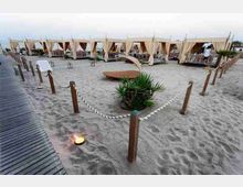 Area lounge su spiaggia sabbiosa con passerella in legno e perimetro delimitato da pali e corde. Sullo sfondo ci sono cabine con tende chiare e lettini rialzati, con piante ornamentali e piccoli bracieri a terra.