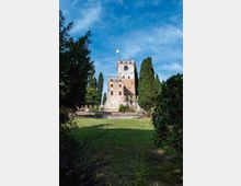 Castello medievale in pietra e mattoni con torre merlata e bandiera in cima, visto da un prato circondato da alberi e cipressi. Il maniero è su un lieve rialzo con un basso muro in pietra, sotto un cielo parzialmente nuvoloso.