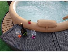 Vasca idromassaggio rotonda con bordo imbottito beige e acqua in movimento, affiancata da una pedana curva in legno e prato sintetico. In primo piano, un tavolino in rattan con una bottiglia e due calici, con una rosa appoggiata sul bordo della vasca.