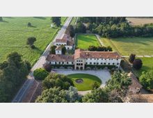 Veduta aerea di una grande villa rurale con edifici in muratura e tetti in coppi, disposti a U attorno a un cortile ghiaioso e a un prato semicircolare con fontana centrale. Intorno si estendono campi coltivati, alberi e una strada rettilinea che costeggia il complesso.