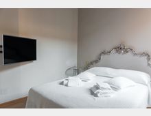Camera da letto minimalista con letto matrimoniale rifatto e biancheria bianca, testiera decorata in stile barocco e asciugamani piegati sul copriletto. Sulla parete laterale è montata una TV a schermo piatto e accanto al letto c’è un piccolo tavolino rotondo in vetro.