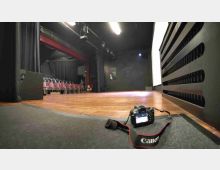 Interno di un piccolo teatro con palco in legno, quinte e tendaggi neri e una fila di sedie rosse sul lato sinistro. In primo piano si vede una fotocamera appoggiata sul pavimento vicino al bordo del palco, con luci e struttura scenica visibili in alto.