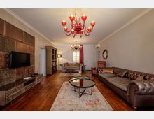 Soggiorno ampio con pavimento in legno, grandi divani in pelle, tappeti chiari e tavolini rotondi con piano in vetro. Sulla parete sinistra ci sono TV e mobile basso decorato, mentre al soffitto pendono lampadari rossi; sul fondo una finestra con tende e una porta.