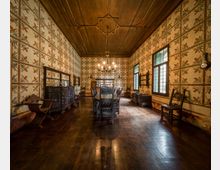 Ampia sala da pranzo storica con pavimento e soffitto in legno, pareti rivestite da carta decorativa a motivi floreali e lampadario centrale. Al centro un lungo tavolo con sedie intagliate, affiancato da credenze e mobili d’epoca, con grandi finestre a vetri che illuminano l’ambiente.