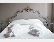 Camera da letto con grande letto matrimoniale rifatto con copriletto bianco, asciugamani piegati e testiera decorativa in stile barocco argentato. Ai lati del letto ci sono due piccoli tavolini rotondi in vetro con supporto metallico, contro una parete grigio chiaro.