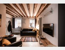 Soggiorno moderno con soffitto a travi in legno a vista, divano nero e tappeto colorato al centro. Due finestre con inferriate e tende illuminano la stanza; sulla parete laterale è montata una TV sopra una mensola in legno rustico.