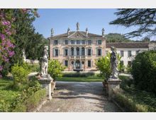 Viale d’accesso in ghiaia che attraversa un giardino alberato e conduce a una villa storica con facciata simmetrica, balconata centrale e statue sul tetto. Ai lati del percorso si vedono due statue su piedistalli, siepi e aiuole fiorite.