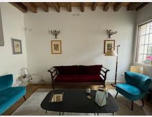 Salotto con soffitto a travi in legno a vista, pareti bianche con quadri e divano scuro al centro, affiancato da due poltrone turchesi. Davanti c’è un tavolino da caffè con oggetti decorativi e, sulla destra, una grande finestra con persiane che illumina l’ambiente.