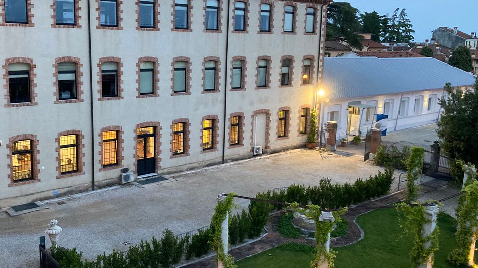 Cortile di un complesso edificio in stile industriale con facciata chiara e finestre incorniciate in mattoni, con pannelli solari sul tetto. In primo piano si vede un giardino curato con prato, siepi e pergolato su colonne, accanto a un vialetto e a un ingresso illuminato verso un’ala bassa.