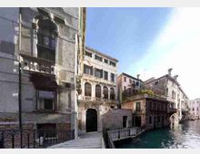 Veduta di un canale urbano con acqua turchese, fiancheggiato da edifici storici in pietra e intonaco con finestre ad arco, balconi e porticati. In primo piano si vede una piccola passerella con ringhiera lungo la riva e una facciata con arco decorativo.