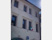 Facciata di un edificio a due piani intonacato di bianco, con grandi finestre rettangolari e un piccolo balcone con bandiera italiana e striscione. In basso si vede un cartello di parcheggio su strada, con cielo sereno sullo sfondo.