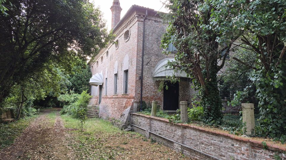 Edificio storico in mattoni rossi con piccole finestre circolari e coperture ad arco sugli ingressi, affacciato su un sentiero sterrato. L’area è circondata da vegetazione fitta e alberi, con un basso muretto in mattoni lungo il lato destro.
