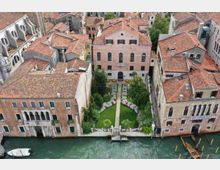 Vista dall’alto di palazzi storici con tetti in coppi rossi affacciati su un canale, con un giardino interno rettangolare tra gli edifici. Il giardino ha un vialetto centrale, prato e aiuole fiorite simmetriche, con un piccolo accesso sul canale.