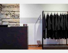 Interno di un negozio con parete in pietra a vista e parete bianca, pavimento in cotto e un bancone scuro. A destra c’è un espositore in metallo con cappotti neri appesi su grucce.