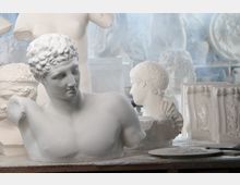 Interno di un laboratorio di scultura con gessi bianchi: in primo piano un busto classico appoggiato su un tavolo, sullo sfondo teste e calchi decorativi allineati su ripiani. L’ambiente appare luminoso, con superfici chiare e attrezzi e materiali da modellazione sul piano di lavoro.