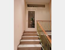 Vano scale interno con gradini in marmo chiaro e alzate effetto legno, affiancato da una ringhiera verde con corrimano in legno. In cima alle scale c’è una porta bianca con pannello in vetro decorato a motivo floreale e una finestra orizzontale sopra la porta.