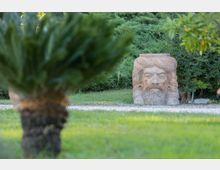 Giardino con prato verde e vegetazione fitta sullo sfondo, attraversato da un vialetto di ghiaia. In fondo si vede una grande scultura in pietra raffigurante una testa barbuta, con una pianta in primo piano sfocata.