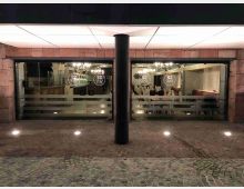 Ingresso di un locale moderno con ampia facciata vetrata e montanti scuri, sotto una pensilina illuminata sostenuta da una colonna centrale. Davanti c’è un’area pavimentata in pietra con luci a incasso, mentre all’interno si vedono tavoli, sedie e lampadari.