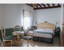 Camera da letto in stile classico con letto matrimoniale e testiera decorata, travi in legno a vista sul soffitto e pareti bianche con fascia verde. Una finestra con tende leggere illumina la stanza, con tavolino rotondo e sedie imbottite vicino al letto.