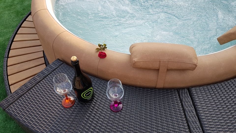 Vasca idromassaggio rotonda con bordo imbottito beige e acqua in movimento, affiancata da una pedana curva in legno e prato sintetico. In primo piano, un tavolino in rattan con una bottiglia e due calici, con una rosa appoggiata sul bordo della vasca.