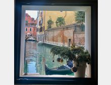 Vista di un canale di Venezia attraverso una finestra, con una gondola ormeggiata in primo piano. Sullo sfondo, edifici storici dai colori vivaci, un ponte ad arco in mattoni e una decorazione natalizia posta sul davanzale della finestra.