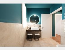 Bagno moderno con pareti verde petrolio e rivestimento in piastrelle beige, lavabo rettangolare su piano in legno scuro e grande specchio rotondo retroilluminato. Accanto a una finestra luminosa ci sono due sgabelli a rete sotto il piano e una porta bianca aperta verso un corridoio con pavimento in legno.