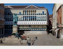 Piazza lastricata con un grande monumento in bronzo su piedistallo, con statua e scultura di leone alato alla base. Sullo sfondo si vede un edificio moderno a più piani con facciata a griglia di finestre e persiane, affiancato da palazzi storici con portici e negozi lungo la strada.