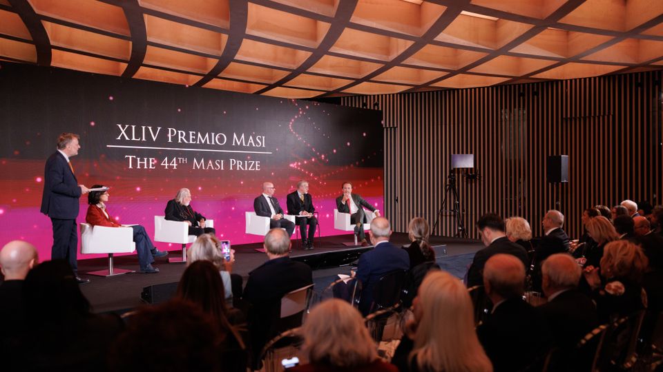 Evento in una sala conferenze moderna con soffitto in legno a griglia e pareti con rivestimento verticale a doghe. Sul palco, uno sfondo luminoso con la scritta "XLIV Premio Masi - The 44th Masi Prize" e diverse persone sedute su poltrone bianche.
