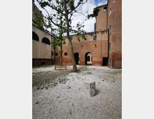 Cortile interno in ghiaia circondato da edifici in mattoni, con un grande albero al centro, panchina in legno e vialetto pavimentato. Sullo sfondo si vedono archi di passaggio e finestre lungo le facciate.