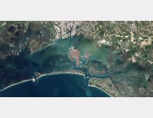 Vista satellitare di una grande laguna costiera con canali e isole, collegata al mare da più bocche tra sottili strisce di terra e diga foranea. Attorno si vedono aree urbane compatte, zone portuali e ampie distese di campi e paludi.