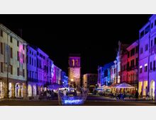 Piazza cittadina di notte con edifici storici ai lati illuminati da luci colorate e portici al piano terra con dehors e ombrelloni. Al centro si vede una torre con grande orologio e arco di passaggio, con un’installazione luminosa in primo piano.