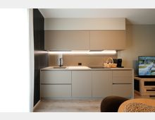 Cucina moderna lineare con mobili grigio opaco, pensili sospesi e illuminazione a LED sotto pensile. Piano di lavoro chiaro con lavello e piano cottura a induzione, pareti neutre e pavimento effetto legno; a destra si intravede un mobile TV in legno.