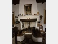 Interno rustico con grande camino in muratura bianca e focolare in pietra, con alari e attrezzi da camino. Sopra la mensola sono esposti piccoli vasi e oggetti decorativi, con un quadro incorniciato al centro e varie stampe e utensili appesi alle pareti laterali.