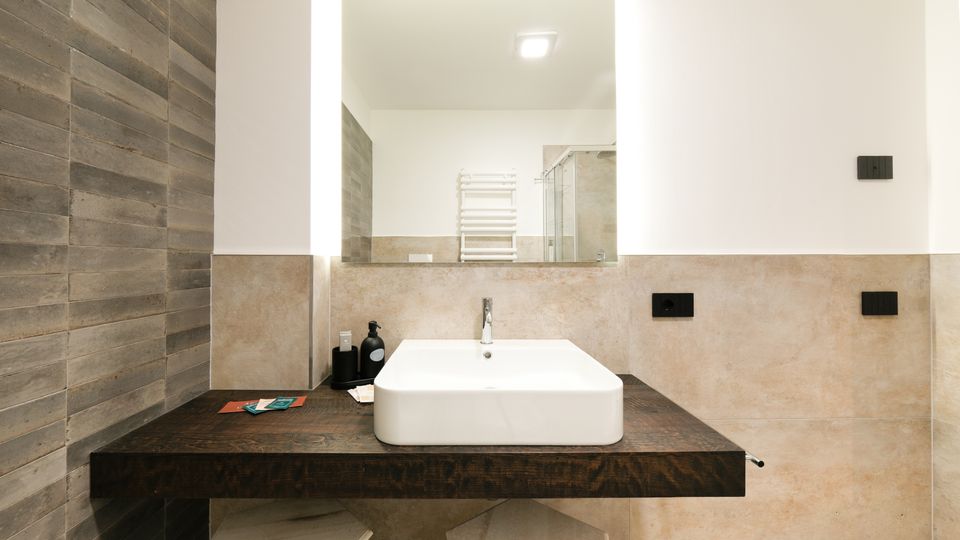 Bagno moderno con lavabo rettangolare in ceramica su mensola in legno scuro, grande specchio e pareti rivestite in piastrelle beige e effetto legno. Sullo sfondo si vede un termoarredo bianco e una cabina doccia in vetro.