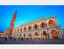 Facciata della Basilica Palladiana in una grande piazza, con logge a due livelli di arcate illuminate e tetto verde. A sinistra si innalza una torre in mattoni con orologio, sullo sfondo di cielo azzurro.