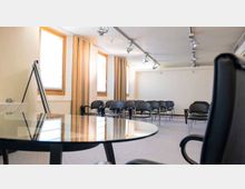 Sala riunioni con file di sedie nere disposte frontalmente verso una parete bianca, illuminata da finestre laterali con tende beige. Sul soffitto sono presenti faretti su binario e in primo piano si vede un tavolo rotondo in vetro.