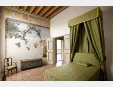 Camera da letto storica con soffitto a travi in legno, grande letto a baldacchino con tende verdi e pavimento in terrazzo. Sulla parete è visibile un affresco parzialmente conservato e una porta aperta conduce a un balcone, con sedia e cassapanca lungo il muro.