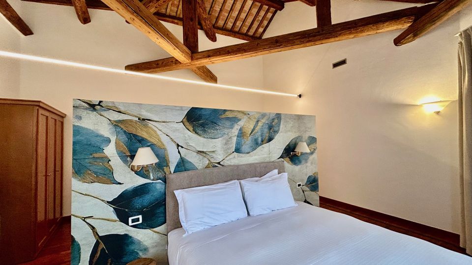 Camera da letto con soffitto alto e travi in legno a vista, pareti chiare e pavimento in legno. Letto matrimoniale con testiera imbottita davanti a una grande carta da parati con foglie blu, applique ai lati e armadio in legno.